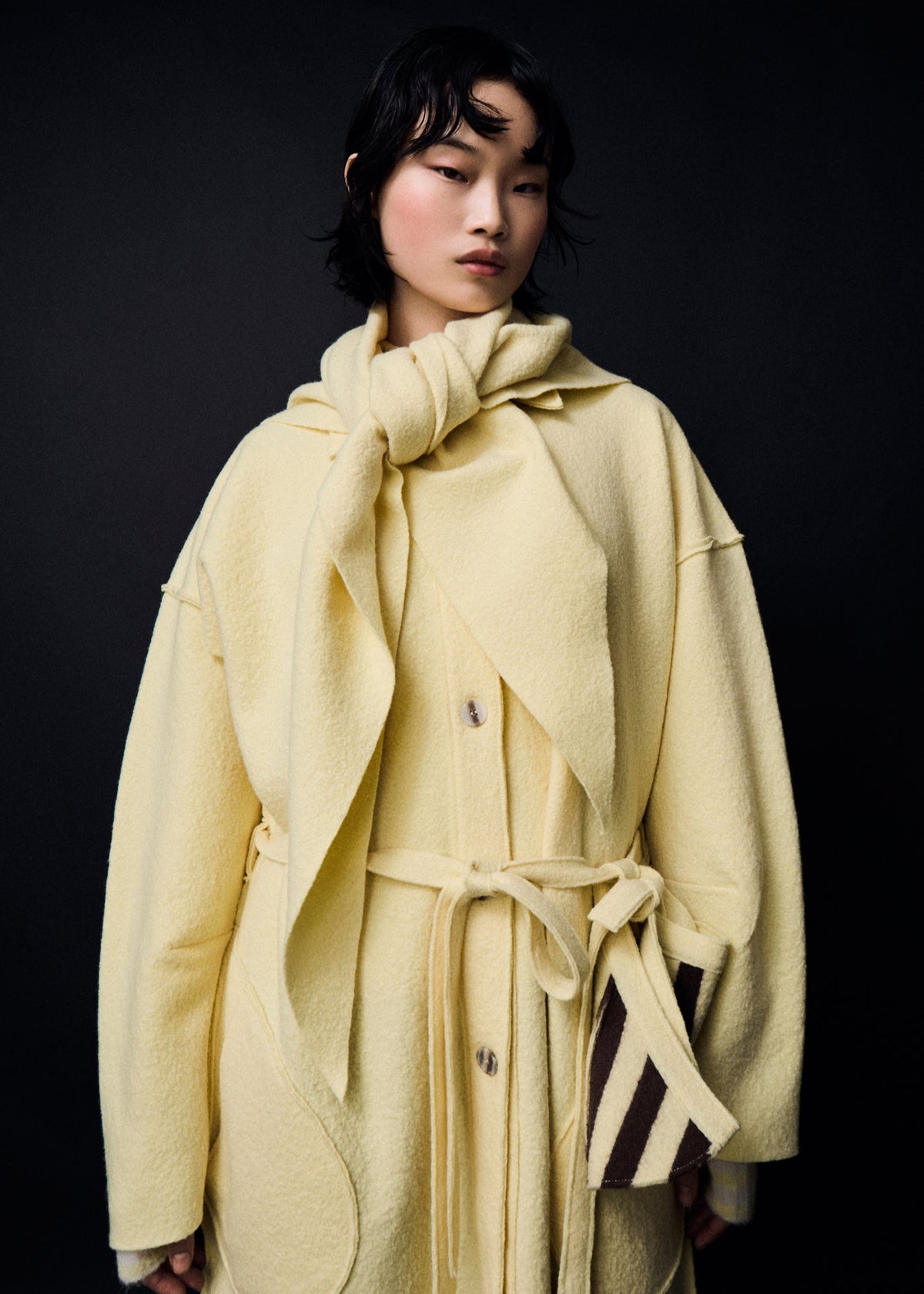 Person wearing a light yellow coat with a matching scarf against a black background