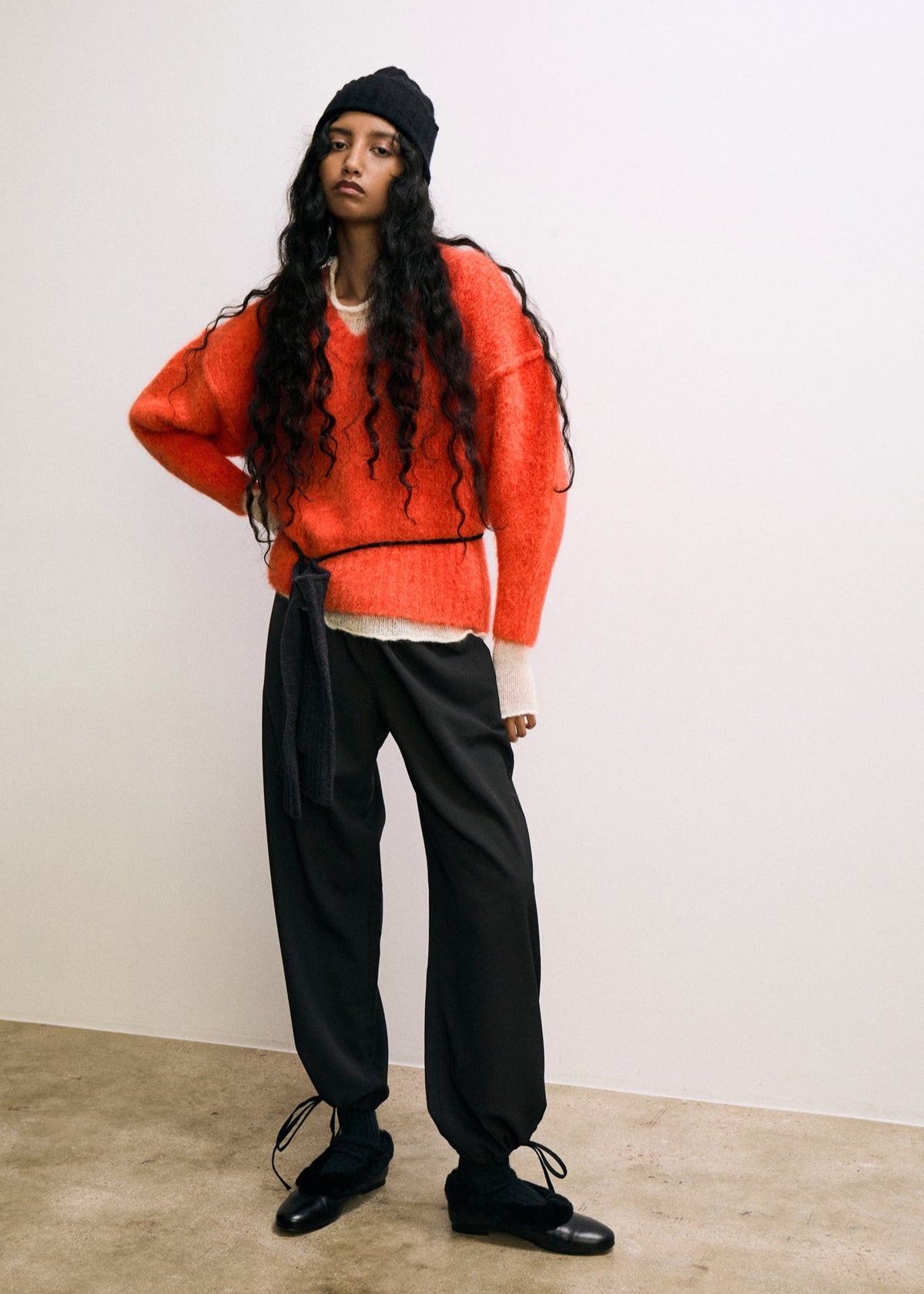 Person wearing a bright orange sweater, black pants, and a black beanie against a white background