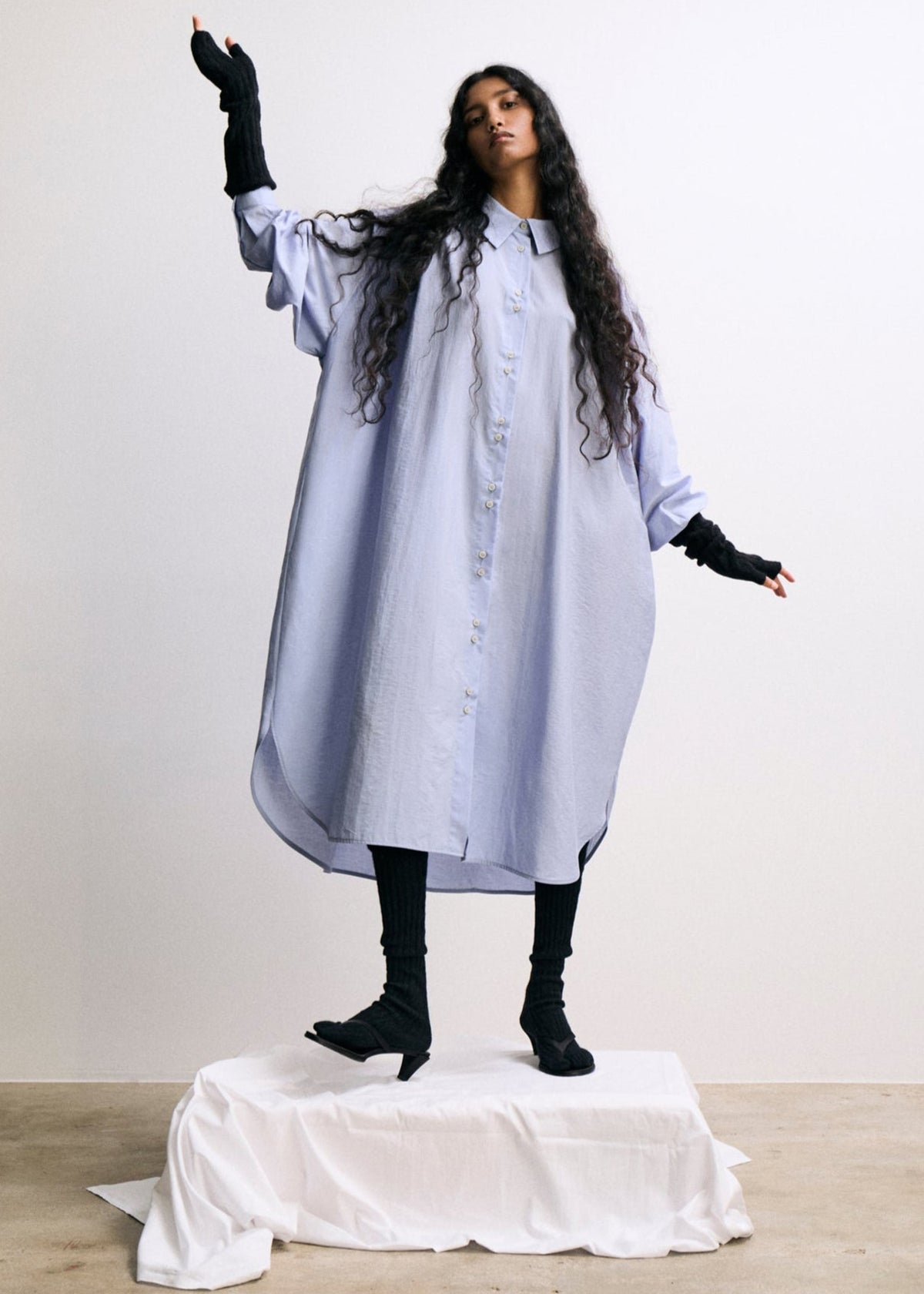 Person wearing a long light blue dress with black gloves and shoes on a white platform against a plain background
