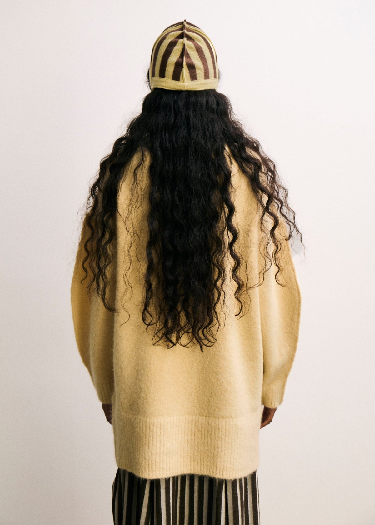 Person wearing a yellow sweater and striped beanie with long, wavy hair on a plain background