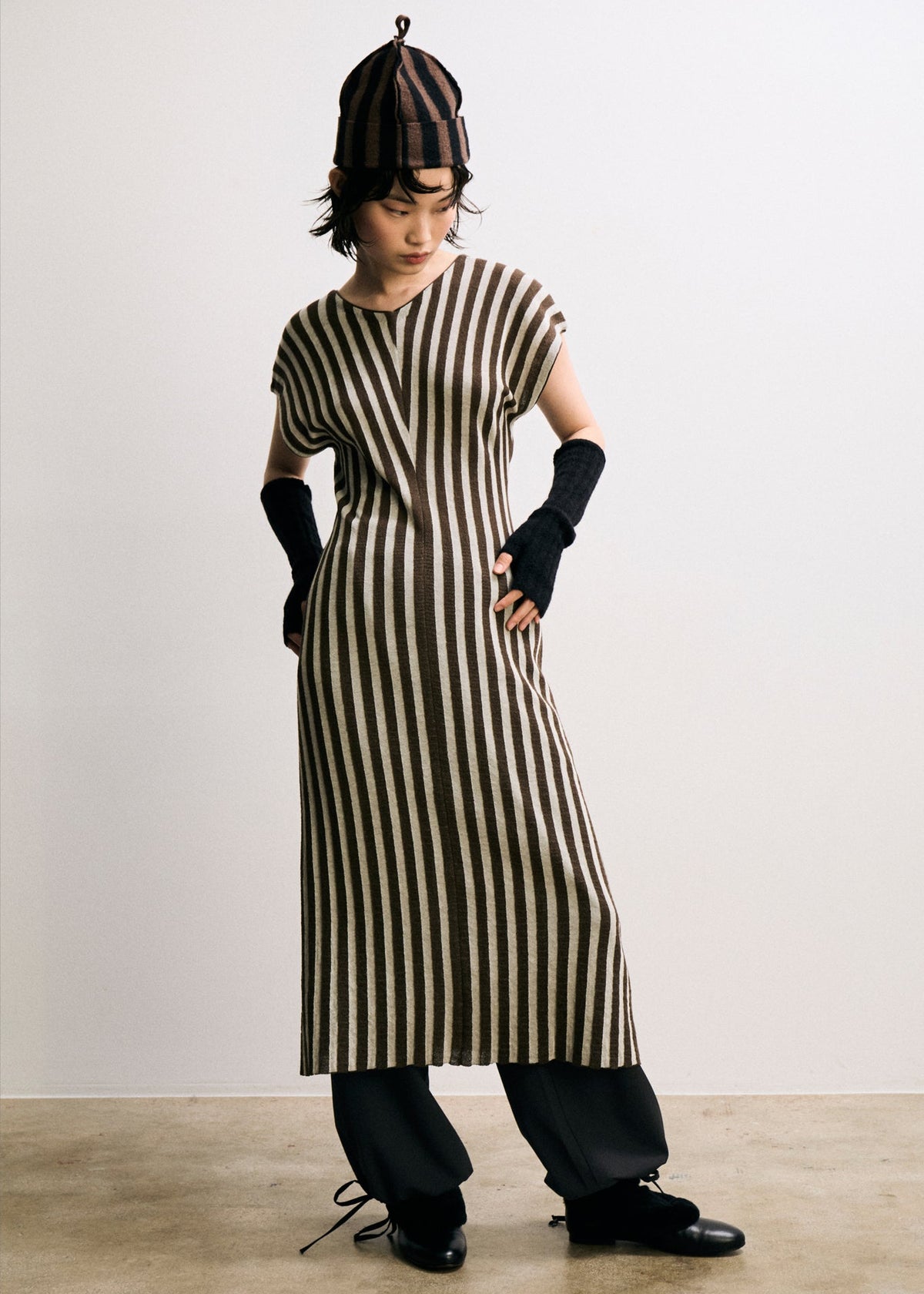 Person wearing a long brown and white striped dress with a hat and gloves on a plain background